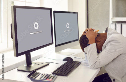 Unhappy, sad businesswoman or professional accountant sitting in front of multiple desktop computers, holding head in despair, waiting for new operating system to load or software update to install