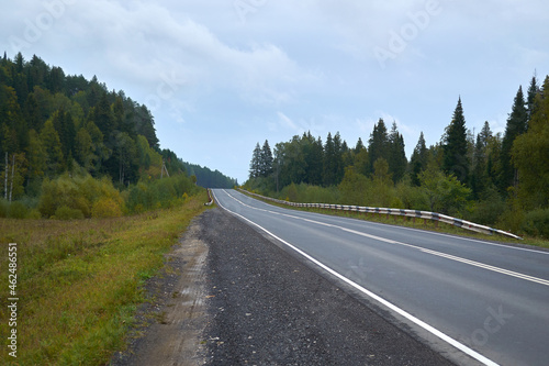 A shallow two-lane highway in Russia in the Urals, passing through the forest. Landscape shot in cloudy daytime. © Slava