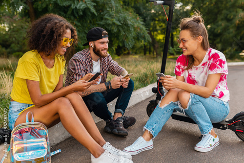 happy young company of smiling friends sitting in park on grass with electric kick scooter