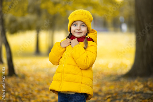 Beautiful outdoor autumn portrait of adorable 4-years old child girl  wearing stylish yellow cap and coat  touching her warm red scarf and smiling at camera.