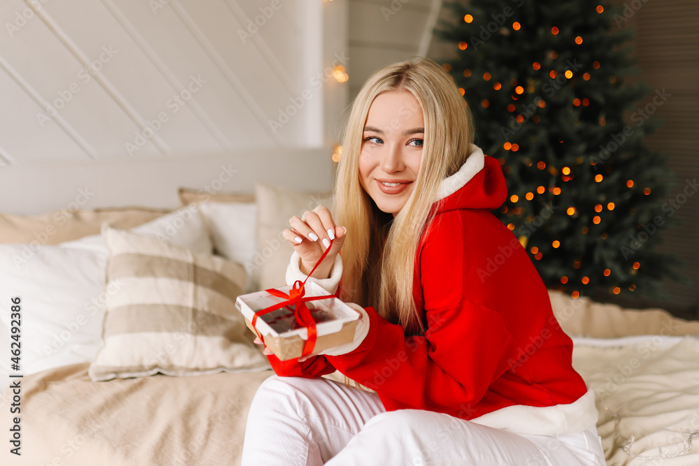 A happy young woman in a red sweater enjoys a gift box present and smiles sitting on the bed in a cozy decorated bedroom in the Christmas holidays at home. Selective focus A happy young woman in a red sweater enjoys a gift box present and smiles sitting on the bed in a cozy decorated bedroom in the Christmas holidays at home. Selective focus