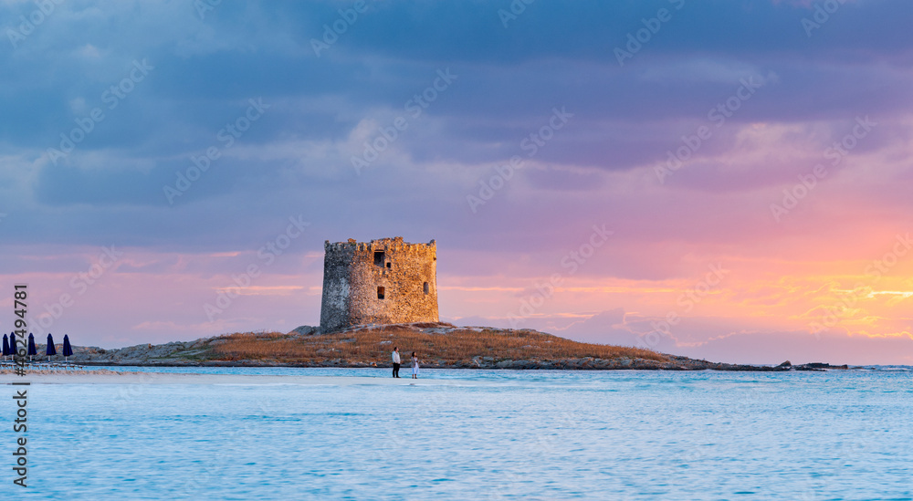 Stunning sunrise over the Aragonese Tower with a couple of tourists enjoying the landscape from La Pelosa Beach, a white sand beach located in Stintino, north-west Sardinia, Italy.