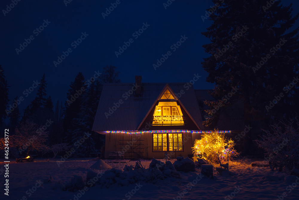 Cute decorated vintage country house home in the middle of forest at night. Tranquil Christmas Eve with snow in garden and light coming from window.