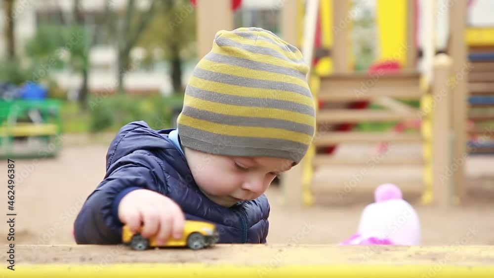 A kid in a warm jacket and hat is playing with a toy car on a playground in the park. Outdoor walks A kid in a warm jacket and hat is playing with a toy car on a playground in the park. Outdoor walks