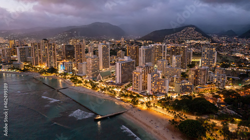 Aerial view of waikiki beach at night in Honolulu, Hawaii. Skyscrapers with lights can be seen with mountains and a sandy beach.