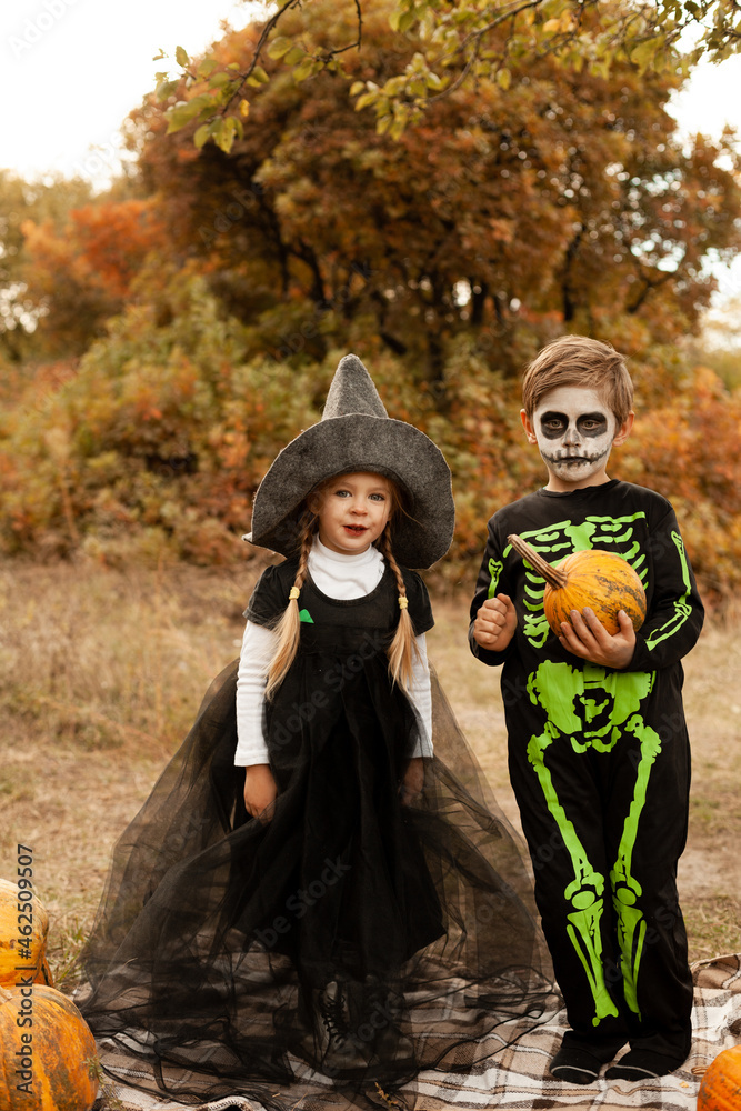 boy and girl dressed as a witch and a skeleton with pumpkins in nature in warm orange colors boy and girl dressed as a witch and a skeleton with pumpkins in nature in warm orange colors