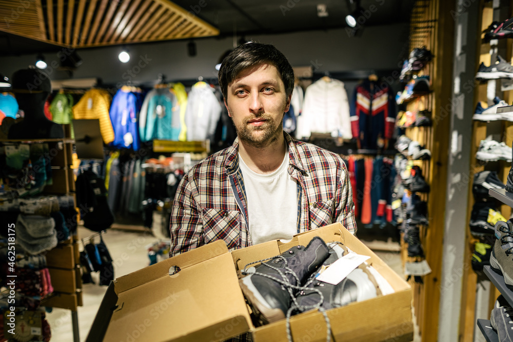 Salesman advises client on choice of hiking shoes in an outdoor store. Male tourist shop owner helping customer try on hiking boots. Young seller confidently presents mountain boot. sell, trade