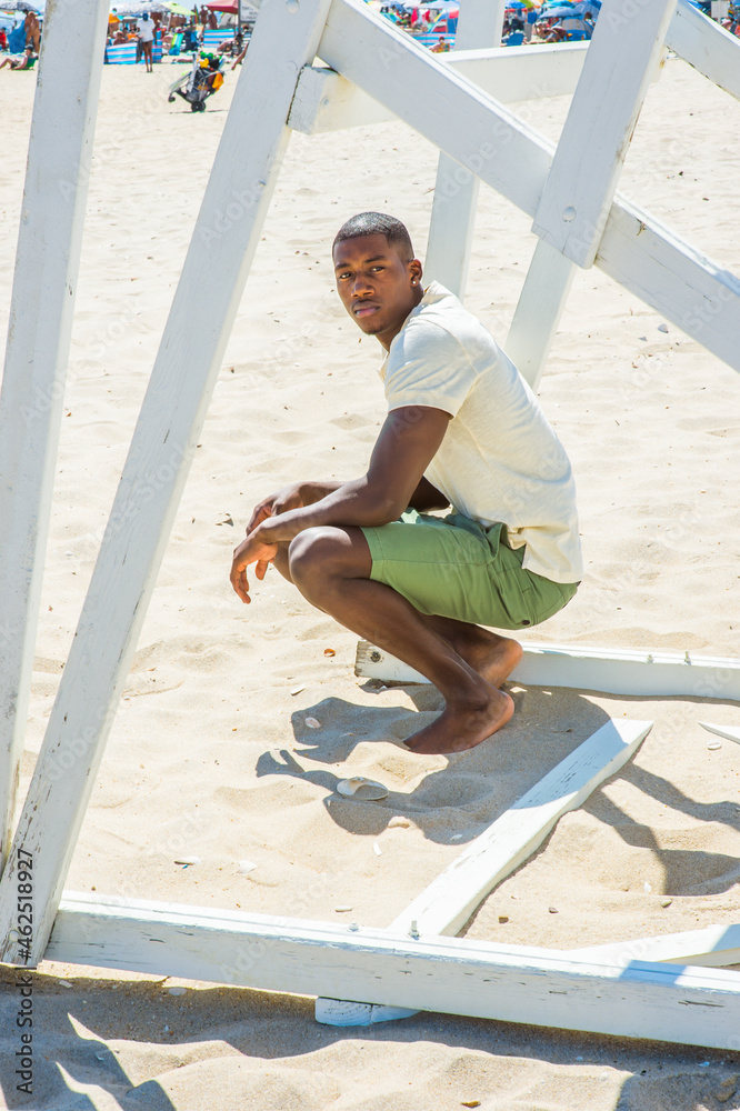 Wearing a white short sleeve shirt, green shorts, barefoot, a young handsome black guy is squatting under a white wooden structure on the beach, looking around.. Many people on background
