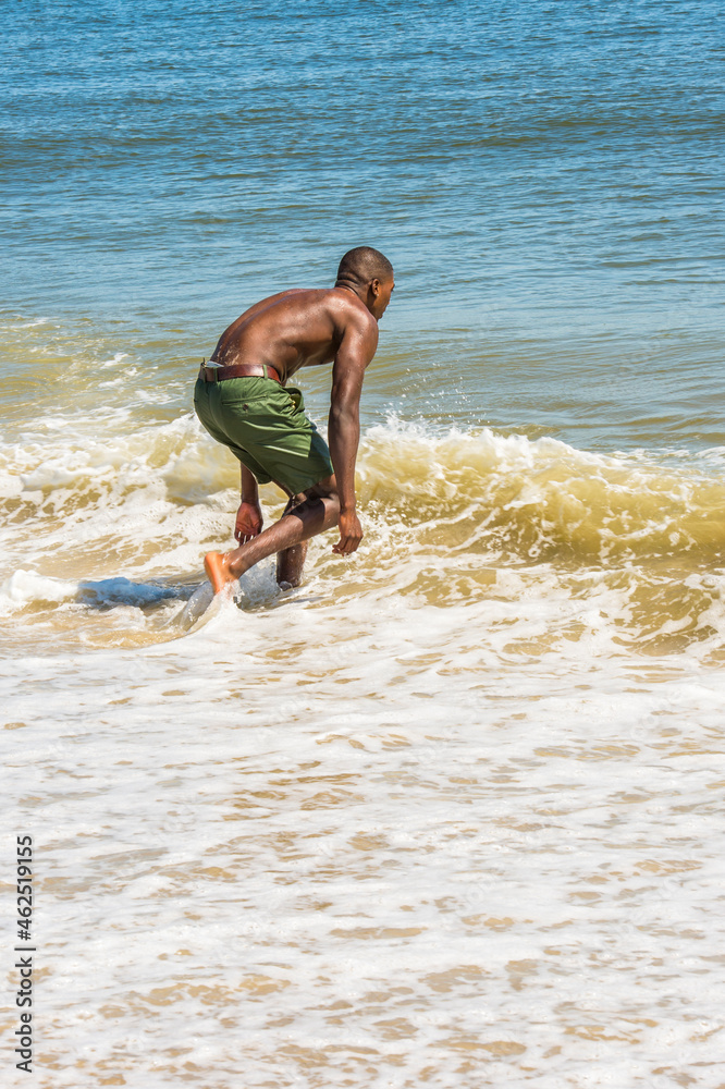 Bending over, a young black man, shirtless, playing with waves of water on beach.