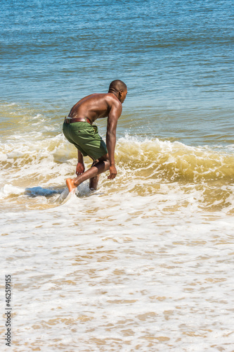 Obraz na płótnie Bending over, a young black man, shirtless, playing with waves of water on beach. w salonie
