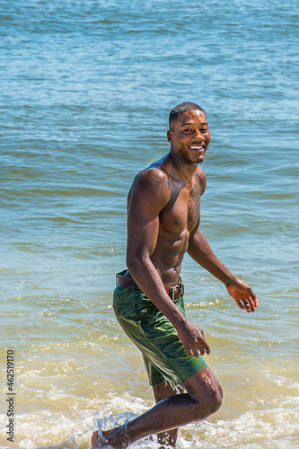 A well built young sexy black guy, half naked and whole body wet,  is walking on the beach, looking at you, smiling.