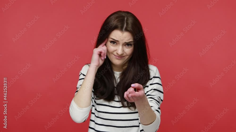 Brunette woman making stupid sign with finger near head and pointing to camera, gesturing bad mind, wearing casual style long sleeve shirt. Indoor studio shot isolated on red background. Brunette woman making stupid sign with finger near head and pointing to camera, gesturing bad mind, wearing casual style long sleeve shirt. Indoor studio shot isolated on red background.