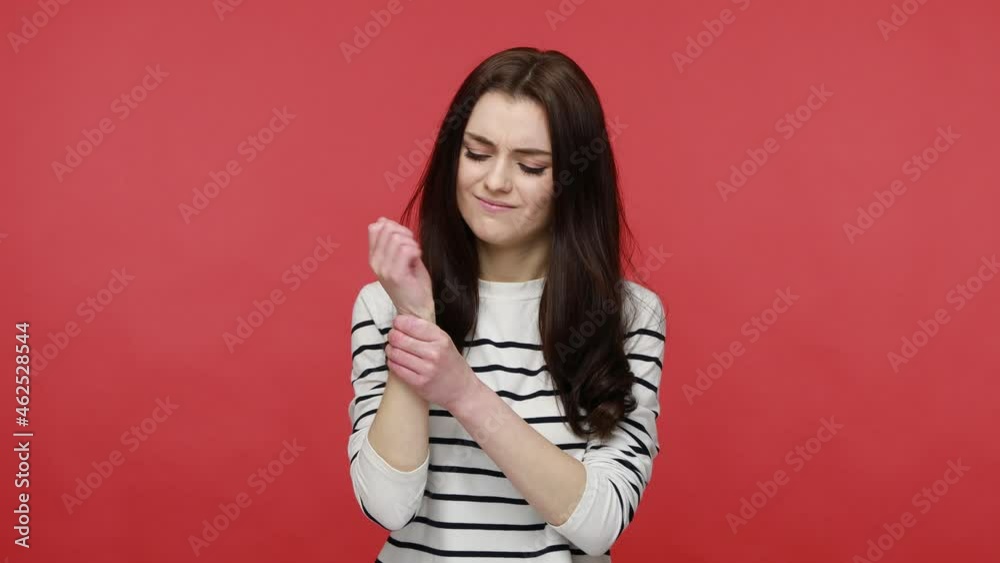 Portrait of woman standing with grimace of pain, massaging sore wrist, suffers hand injury or sprain, wearing casual style long sleeve shirt. Indoor studio shot isolated on red background. Portrait of woman standing with grimace of pain, massaging sore wrist, suffers hand injury or sprain, wearing casual style long sleeve shirt. Indoor studio shot isolated on red background.