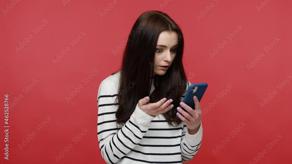 Woman scrolling social media using cell phone, becoming surprised amazed by mobile application, wearing casual style long sleeve shirt. Indoor studio shot isolated on red background.