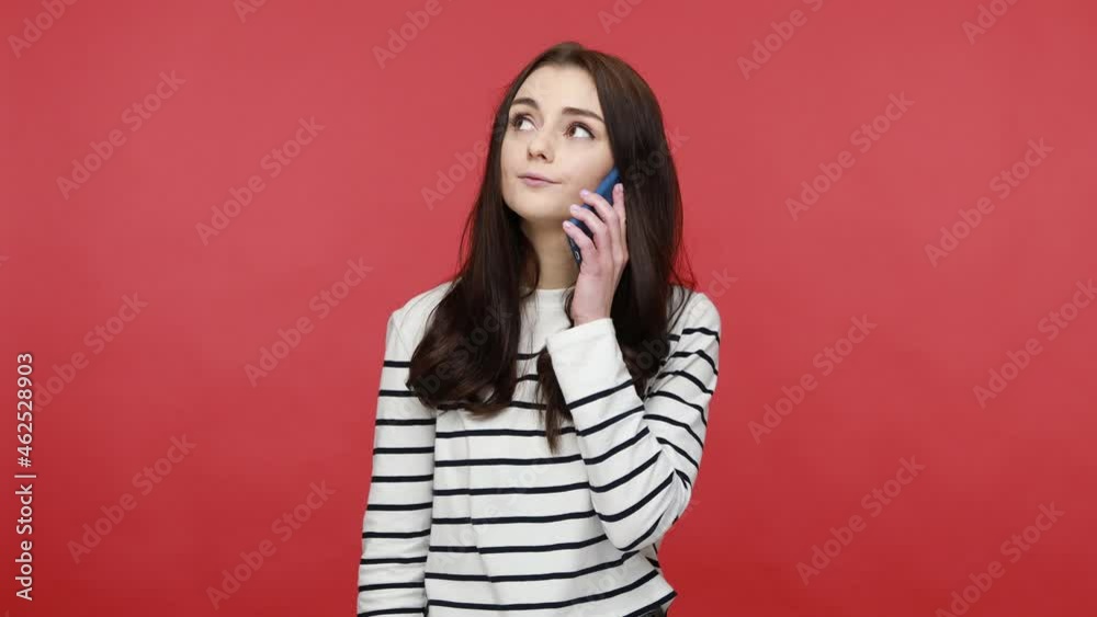 Portrait of woman calling and talking on cellphone, having amazed face after hearing shocking news, wearing casual style long sleeve shirt. Indoor studio shot isolated on red background.