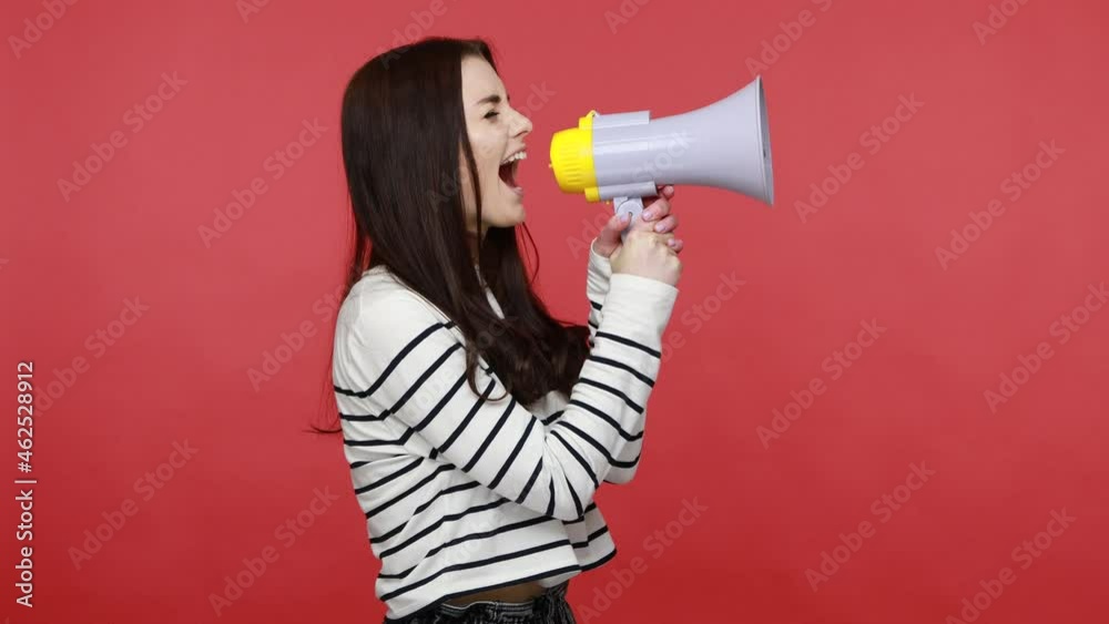 Side view portrait of woman talking on megaphone, proclaiming news, loudly announcing advertisement, wearing casual style long sleeve shirt. Indoor studio shot isolated on red background. Side view portrait of woman talking on megaphone, proclaiming news, loudly announcing advertisement, wearing casual style long sleeve shirt. Indoor studio shot isolated on red background.