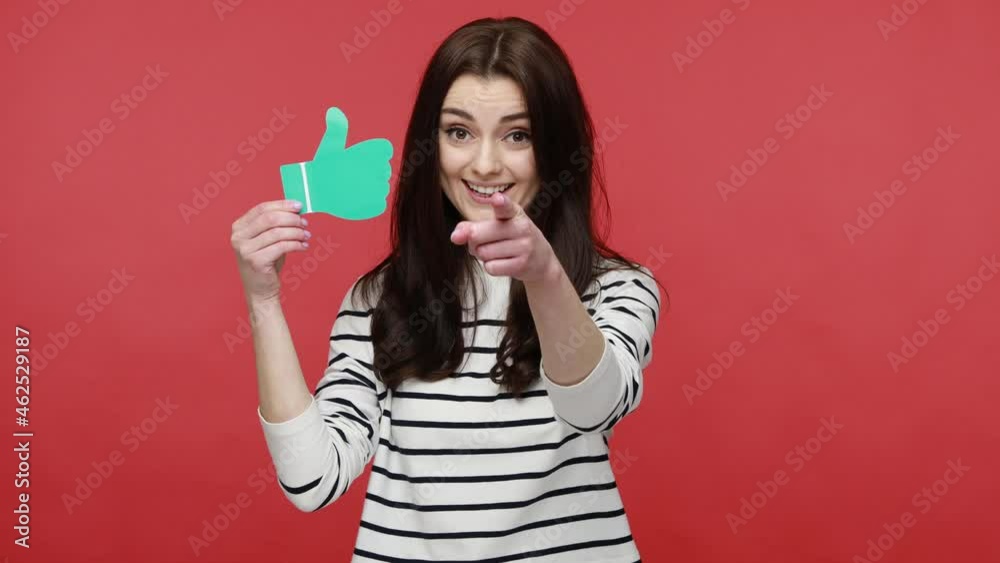 Portrait of pretty woman holding and showing like or thumbs up paper shape sign, pointing to camera, wearing casual style long sleeve shirt. Indoor studio shot isolated on red background. Portrait of pretty woman holding and showing like or thumbs up paper shape sign, pointing to camera, wearing casual style long sleeve shirt. Indoor studio shot isolated on red background.
