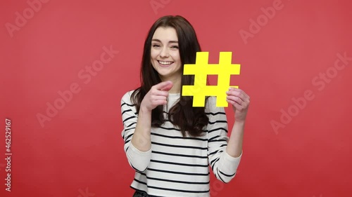 Woman points finger at yellow hashtag symbol, gives recommendation to join forum, popular blog idea, wearing casual style long sleeve shirt. Indoor studio shot isolated on red background.