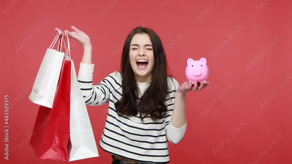 Portrait of positive young woman showing shopping bags and piggy bank, savings money, cashback, wearing casual style long sleeve shirt. Indoor studio shot isolated on red background. Portrait of positive young woman showing shopping bags and piggy bank, savings money, cashback, wearing casual style long sleeve shirt. Indoor studio shot isolated on red background.