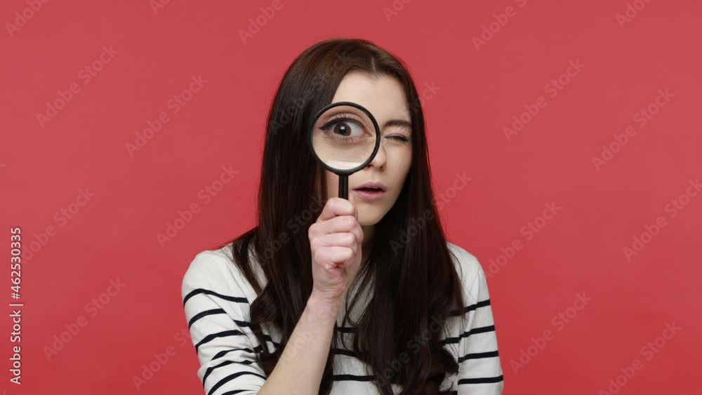 Portrait of woman looks into camera through magnifying glass, inspects something, private detective, wearing casual style long sleeve shirt. Indoor studio shot isolated on red background.