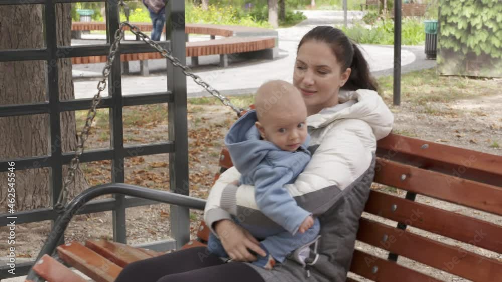 Mother with her child on wooden swinging park bench in a public place, mom with baby boy outdoors. Happy family. High quality 4k footage
