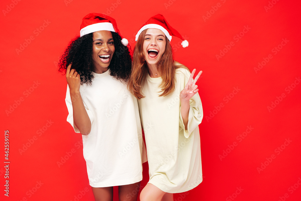 Two beautiful women celebrating New Year.Happy gorgeous female. They having fun at New Year's Eve party.Holiday celebration.Charming models in Santa Claus hats laughing near red wall Two beautiful women celebrating New Year.Happy gorgeous female. They having fun at New Year's Eve party.Holiday celebration.Charming models in Santa Claus hats laughing near red wall