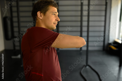 Portrait of handsome guy with short haircut half-turned to camera in sport shirt doing hand stretching, isolated against gym equipment background, doing everyday fitness exercises to become stronger