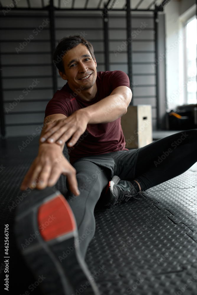 Good-looking slim guy training endurance and flexibility, stretching legs and hands during warming up exercises before intensive workout at gym, sitting on floor against horizontal bars Good-looking slim guy training endurance and flexibility, stretching legs and hands during warming up exercises before intensive workout at gym, sitting on floor against horizontal bars