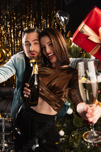happy woman holding glass and bottle of champagne near thrilled man with christmas present on black