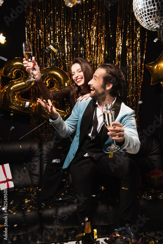 cheerful couple with champagne celebrating new year on sofa near shiny decoration on black