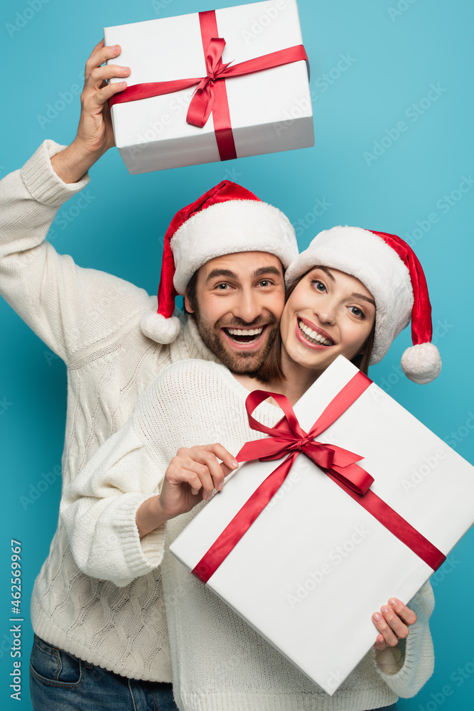 cheerful couple in santa hats and white sweaters holding christmas presents on blue cheerful couple in santa hats and white sweaters holding christmas presents on blue