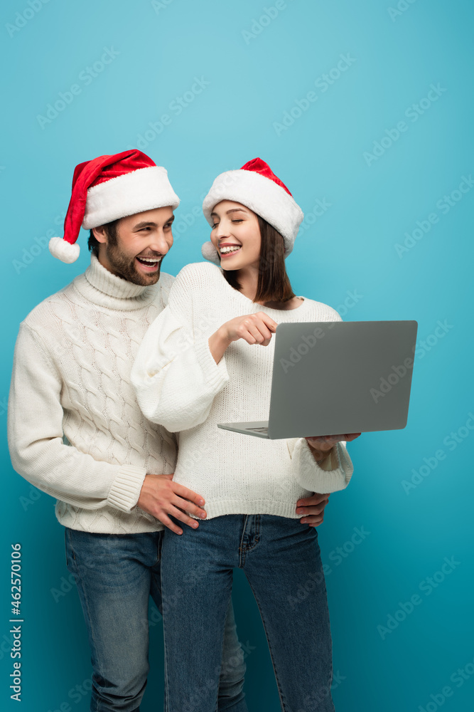 cheerful woman pointing at laptop near smiling man in santa hat on blue