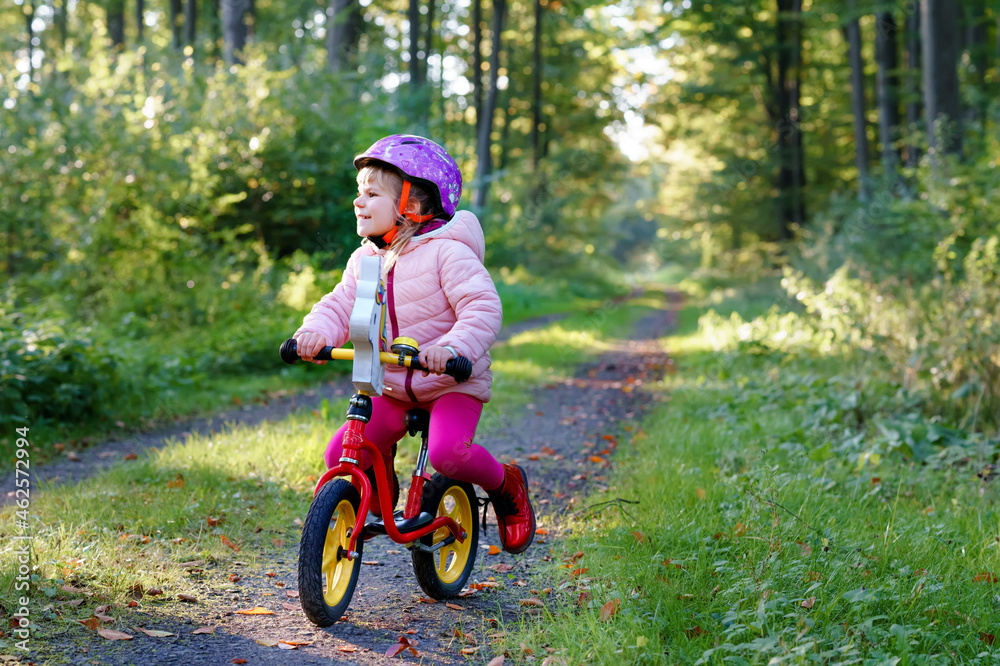 Cute little toddler girl with helmet riding on run balance bike to daycare, playschool or kindergarden. Happy child having fun with learning on learner bicycle. Active kid on cold autumn day outdoors. Cute little toddler girl with helmet riding on run balance bike to daycare, playschool or kindergarden. Happy child having fun with learning on learner bicycle. Active kid on cold autumn day outdoors.