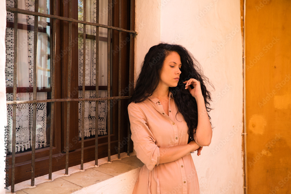 Fototapeta premium beautiful woman with dark curly hair leaning against the wall and the window grille of a typical Mediterranean style house. The woman is serious and sad. Concept expressions.