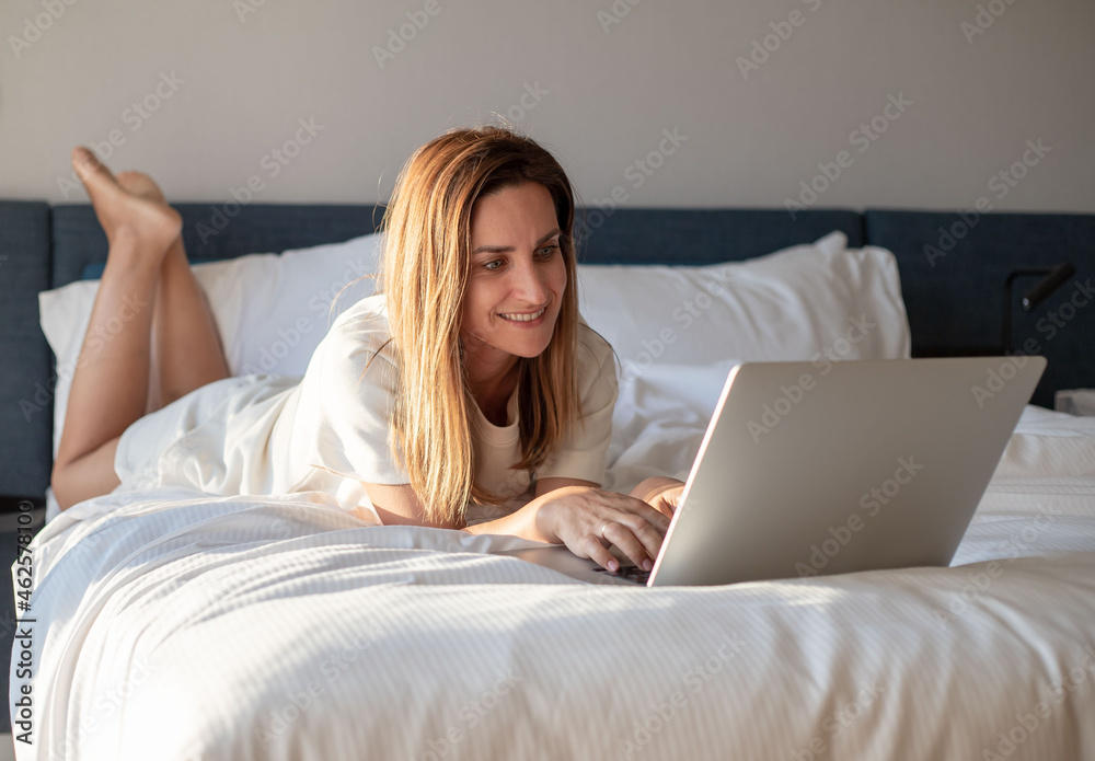Woman lying in bed with laptop smiling. Woman lying in bed with laptop smiling.
