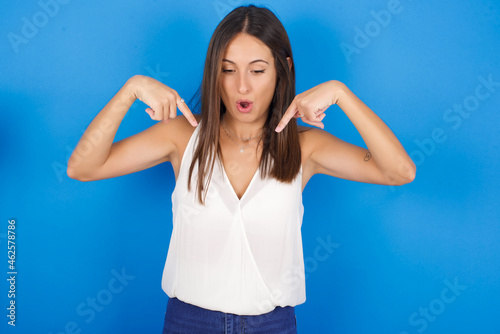 Amazed Young european brunette woman wearing white T-shirt on blue background points down with fore fingers, opens mouth being shocked. Advertisement concept.