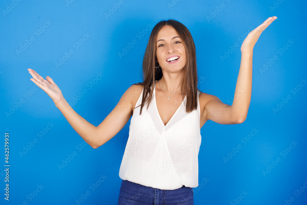 Cheerful Young european brunette woman wearing white T-shirt on blue background making a welcome gesture raising arms over head. Cheerful Young european brunette woman wearing white T-shirt on blue background making a welcome gesture raising arms over head.
