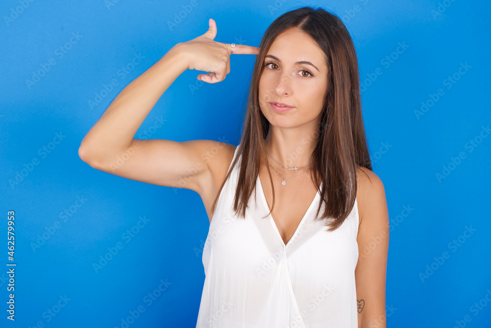 Unhappy Young european brunette woman wearing white T-shirt on blue background curves lips and makes suicide gesture, shoots in temple with hand, tries kill himself. Unhappy Young european brunette woman wearing white T-shirt on blue background curves lips and makes suicide gesture, shoots in temple with hand, tries kill himself.