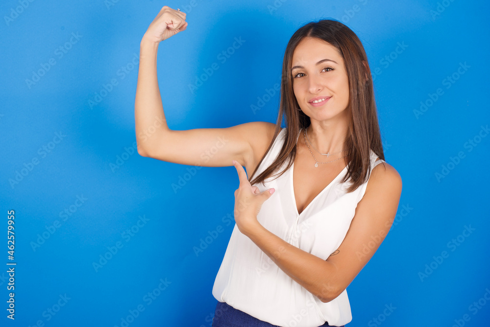 Fototapeta premium Smiling Young european brunette woman wearing white T-shirt on blue background raises hand to show muscles, feels confident in victory, strong and independent.