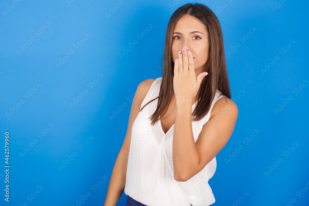 Oh! I think I said it! Close up portrait Young european brunette woman wearing white T-shirt on blue background cover open mouth by hand palm, look at camera with big eyes. Oh! I think I said it! Close up portrait Young european brunette woman wearing white T-shirt on blue background cover open mouth by hand palm, look at camera with big eyes.
