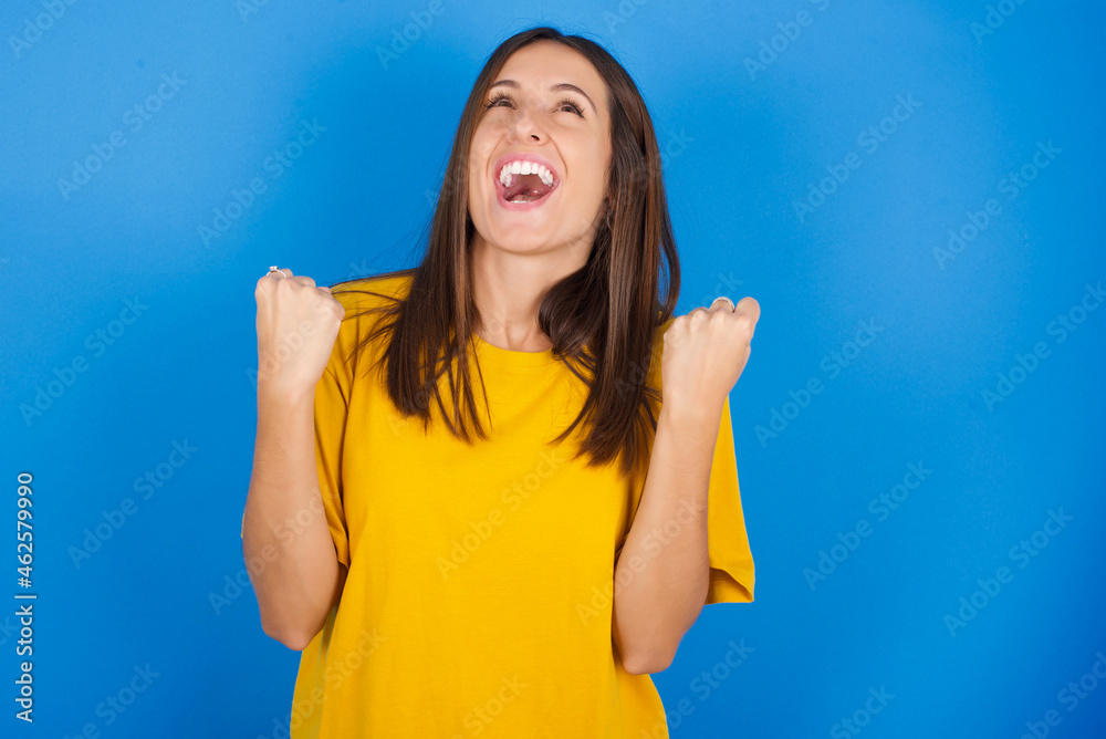Young european brunette woman wearing yellow T-shirt on blue background looks with excitement up, keeps hands raised, notices something unexpected. Young european brunette woman wearing yellow T-shirt on blue background looks with excitement up, keeps hands raised, notices something unexpected.