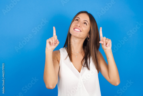 Successful friendly looking Young european brunette woman wearing white T-shirt on blue background exclaiming excitedly, pointing both index fingers up, indicating something.