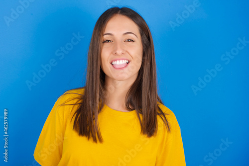 Young european brunette woman wearing yellow T-shirt on blue background with happy and funny face smiling and showing tongue.