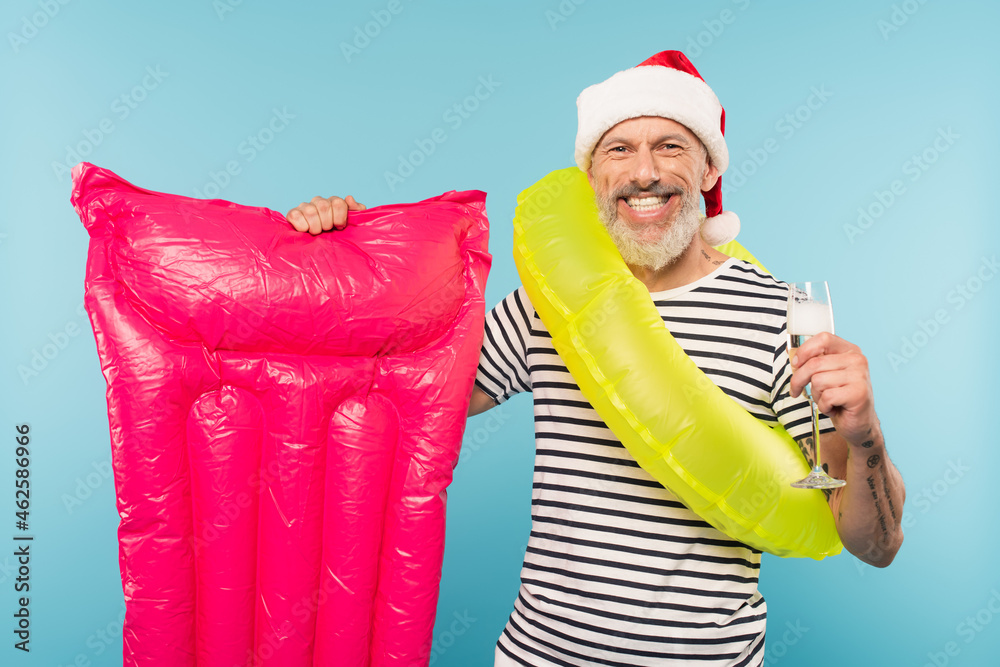 Obraz premium cheerful middle aged man in santa hat and swim ring holding pink inflatable mattress and glass of champagne isolated on blue