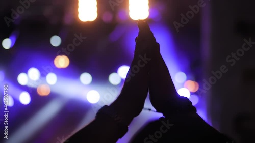 Slow motion, close up: man silhouette partying, cheering, raising hands up and clapping at rock concert in front of stage of nightclub. Bright colorful stage lighting. Nightlife, entertainment concept