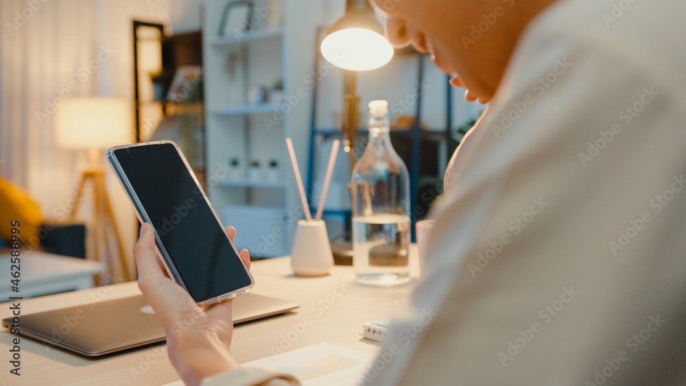 Young Asia lady use smart phone with blank black screen mock up display for advertising text while smart working from home at living room at night. Chroma key technology, Marketing design concept.