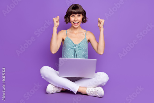Portrait of attractive cheerful lucky girl sitting on floor using laptop rejoicing isolated over bright purple violet color background