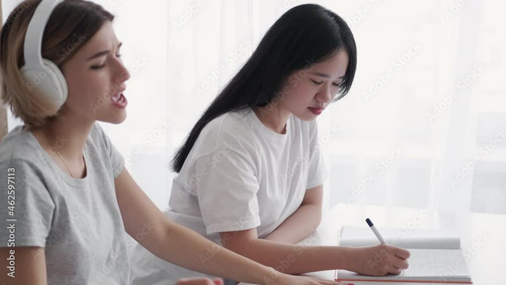 Distracted student. Boring homework. Carefree millennial. Music lover. Mixed race Asian Caucasian girls unmotivated classmate enjoying headphones song singing in class interior. Distracted student. Boring homework. Carefree millennial. Music lover. Mixed race Asian Caucasian girls unmotivated classmate enjoying headphones song singing in class interior.