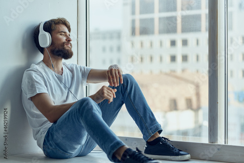 man in white t-shirt sitting near the window in headphones listening to music