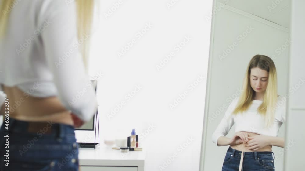 A young woman stands in front of a mirror and measures her waist circumference with a tape measure A young woman stands in front of a mirror and measures her waist circumference with a tape measure
